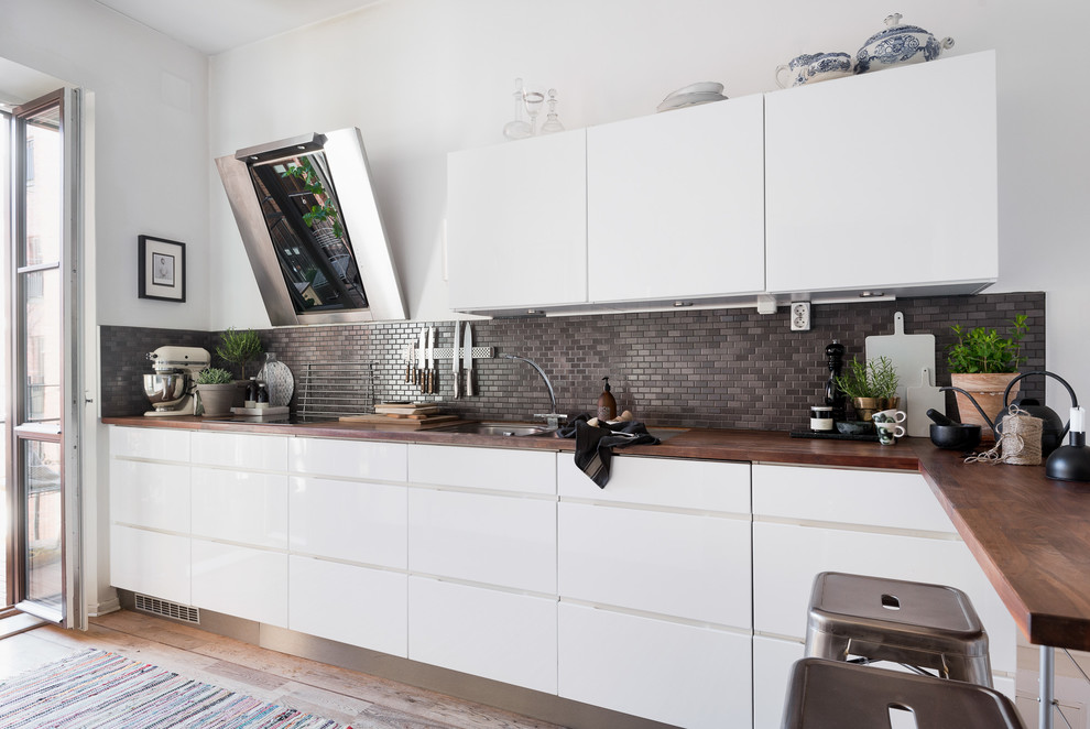 Inredning av ett skandinaviskt mellanstort linjärt kök med öppen planlösning, med släta luckor, vita skåp, träbänkskiva, grått stänkskydd, stänkskydd i mosaik, vita vitvaror, mellanmörkt trägolv och brunt golv