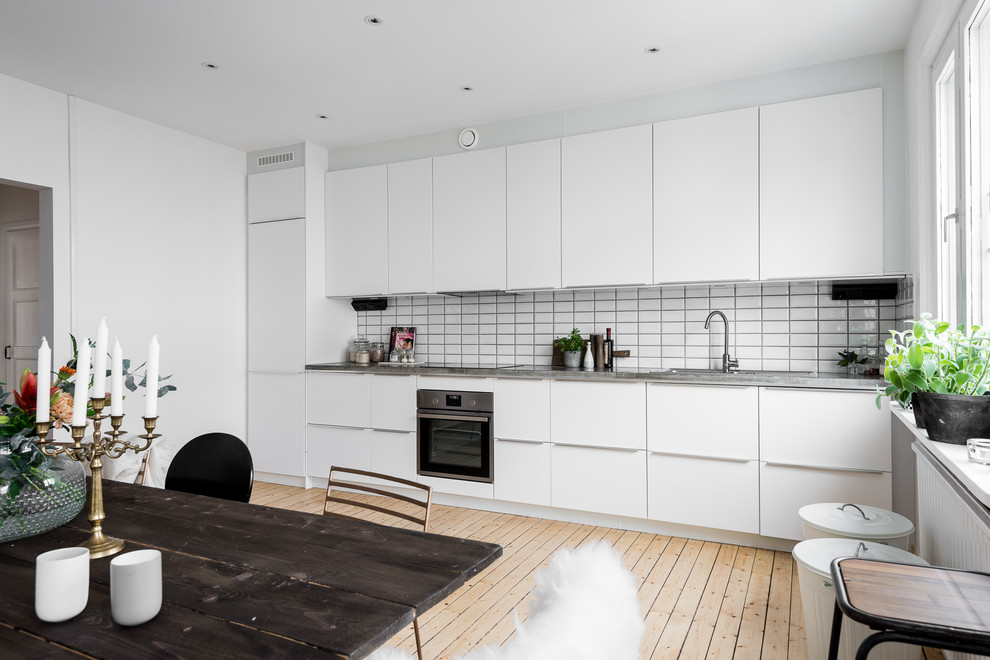 Foto på ett funkis linjärt kök och matrum, med en enkel diskho, släta luckor, vita skåp, bänkskiva i betong, vitt stänkskydd, stänkskydd i keramik, vita vitvaror och ljust trägolv