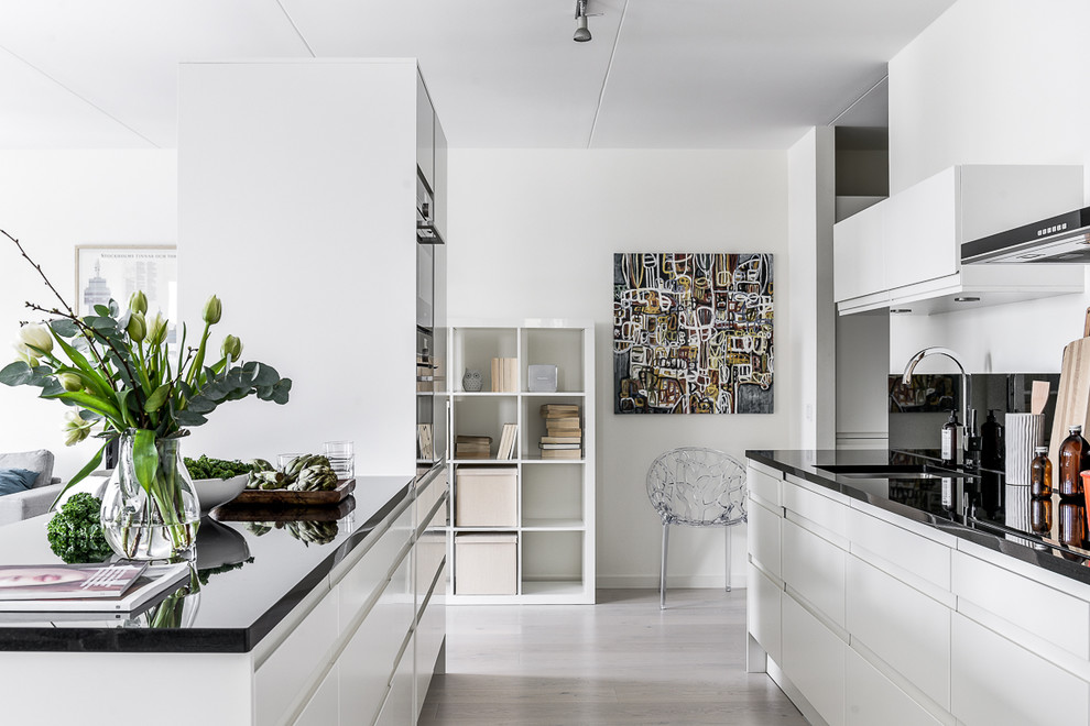 Inspiration for a large modern galley open plan kitchen in Stockholm with a submerged sink, flat-panel cabinets, black splashback, black appliances, light hardwood flooring and an island.