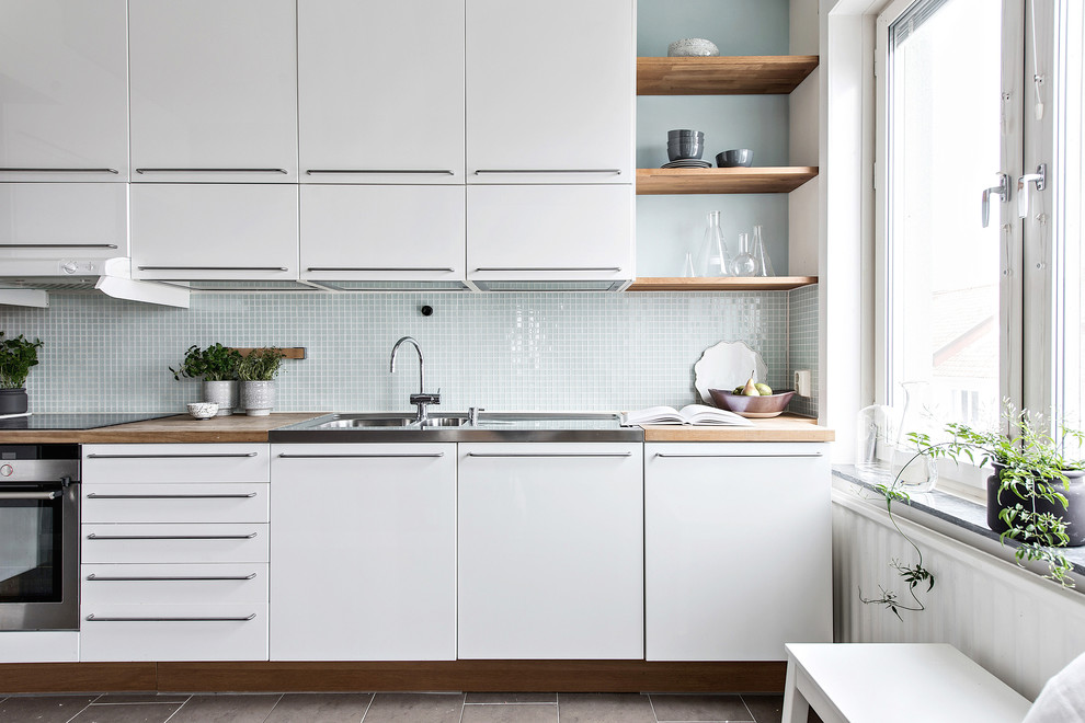 Skandinavisk inredning av ett linjärt kök, med släta luckor, vita skåp, träbänkskiva, grått stänkskydd, stänkskydd i mosaik och brunt golv
