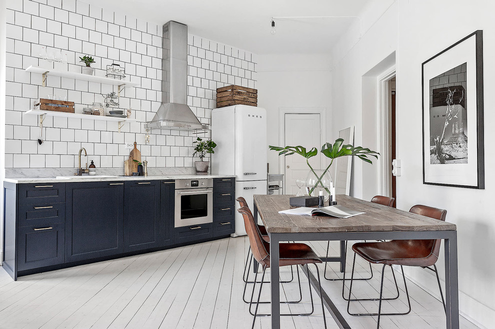 Стильный дизайн: прямая кухня в скандинавском стиле с обеденным столом, накладной мойкой, фасадами с утопленной филенкой, синими фасадами, белым фартуком, белой техникой, деревянным полом и белым полом - последний тренд