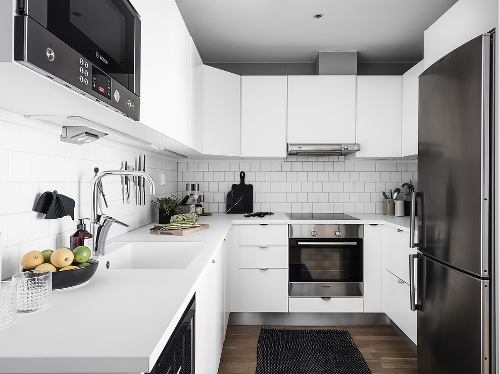 Exempel på ett litet minimalistiskt vit vitt u-kök, med en integrerad diskho, släta luckor, vita skåp, vitt stänkskydd, rostfria vitvaror, mellanmörkt trägolv och brunt golv