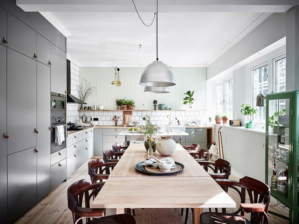 Inspiration for a scandinavian l-shaped kitchen/diner in Gothenburg with a belfast sink, flat-panel cabinets, grey cabinets, wood worktops, white splashback, black appliances, light hardwood flooring, an island and beige floors.