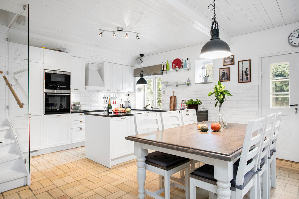 Photo of a large rural u-shaped kitchen/diner in Malmo with recessed-panel cabinets, white cabinets, white splashback, black appliances, brick flooring and an island.