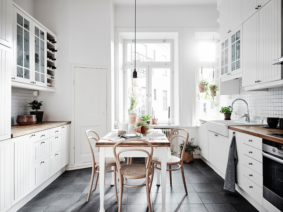 Design ideas for a scandi galley kitchen/diner in Gothenburg with a belfast sink, white cabinets, wood worktops, white splashback, stainless steel appliances, no island and black floors.