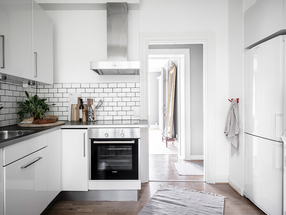 This is an example of a scandinavian u-shaped kitchen in Gothenburg with an integrated sink, flat-panel cabinets, white cabinets, stainless steel worktops, white splashback, ceramic splashback, white appliances, medium hardwood flooring, no island, brown floors and grey worktops.