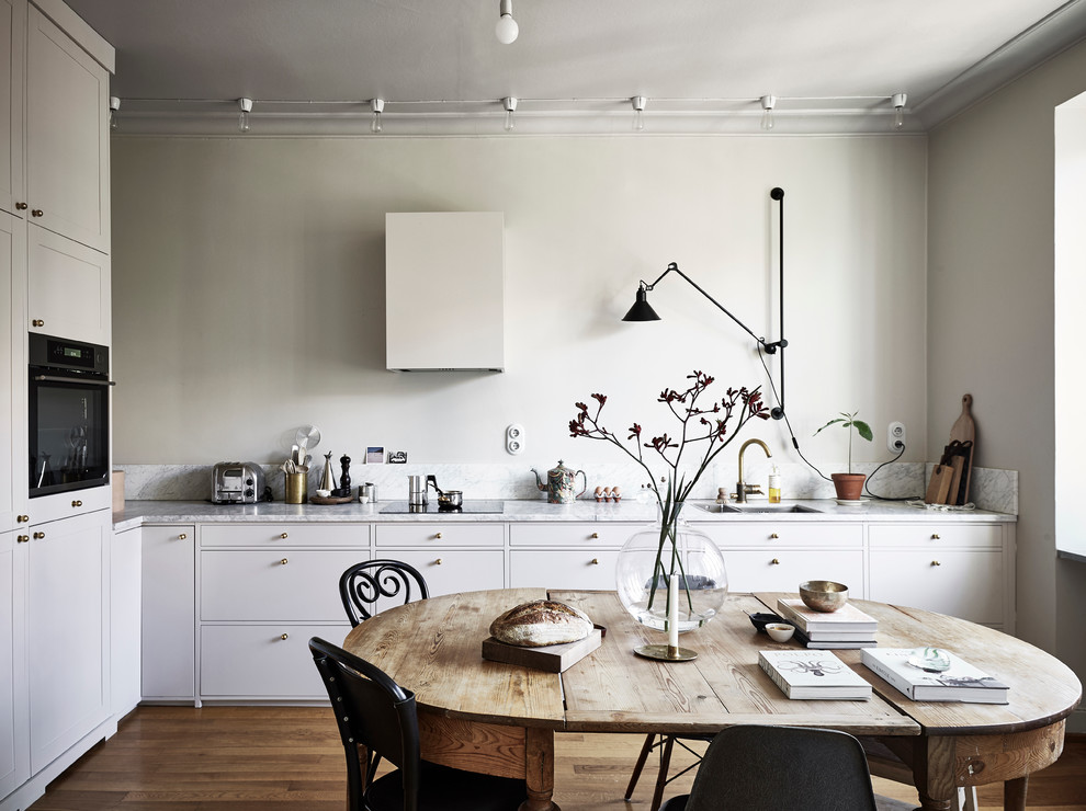 Foto på ett mellanstort nordiskt kök, med en dubbel diskho, släta luckor, vita skåp, marmorbänkskiva, grått stänkskydd, vita vitvaror och mellanmörkt trägolv