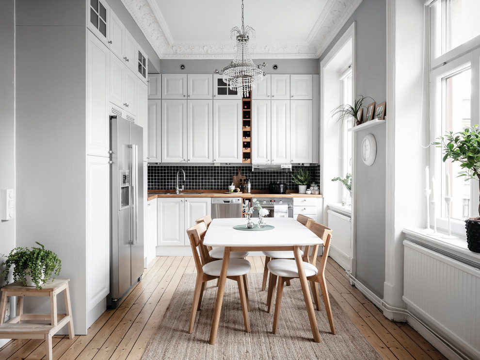 Danish l-shaped medium tone wood floor and brown floor eat-in kitchen photo in Gothenburg with raised-panel cabinets, white cabinets, wood countertops, black backsplash, stainless steel appliances, no island and brown countertops