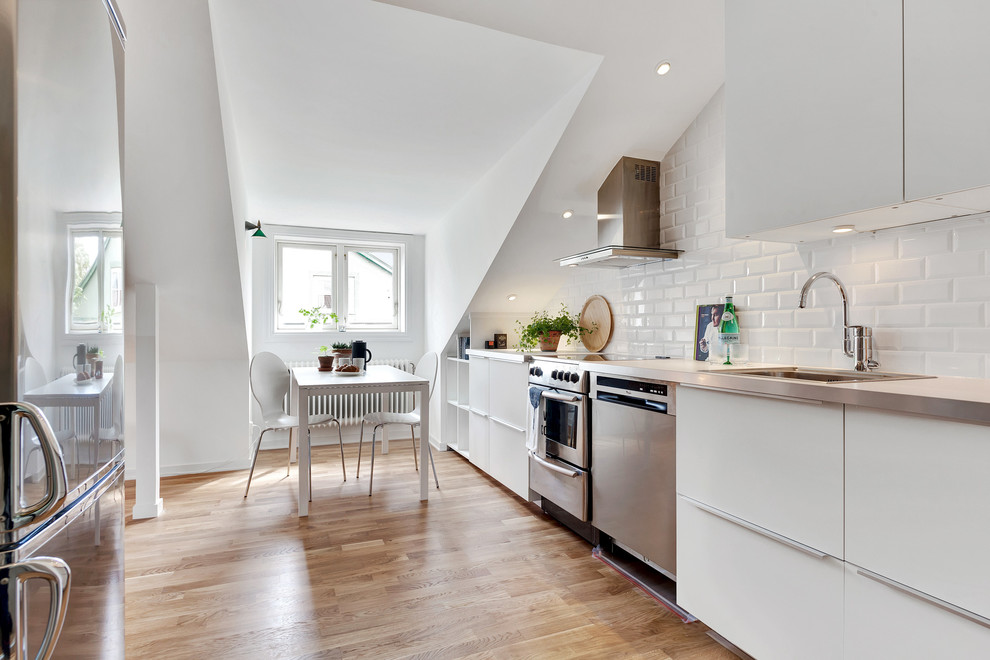 Foto på ett skandinaviskt linjärt kök och matrum, med en nedsänkt diskho, släta luckor, vita skåp, bänkskiva i rostfritt stål, vitt stänkskydd, stänkskydd i tunnelbanekakel, rostfria vitvaror och ljust trägolv