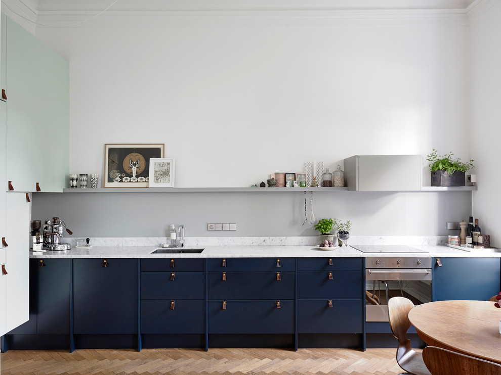 Inredning av ett minimalistiskt mellanstort linjärt kök och matrum, med en undermonterad diskho, blå skåp, grått stänkskydd, rostfria vitvaror, ljust trägolv, släta luckor och marmorbänkskiva