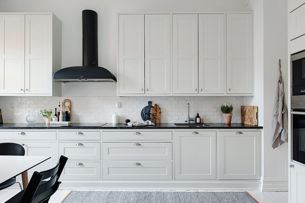 Idéer för att renovera ett avskilt, stort minimalistiskt linjärt kök, med en enkel diskho, vita skåp, granitbänkskiva, vitt stänkskydd, svarta vitvaror, målat trägolv och skåp i shakerstil