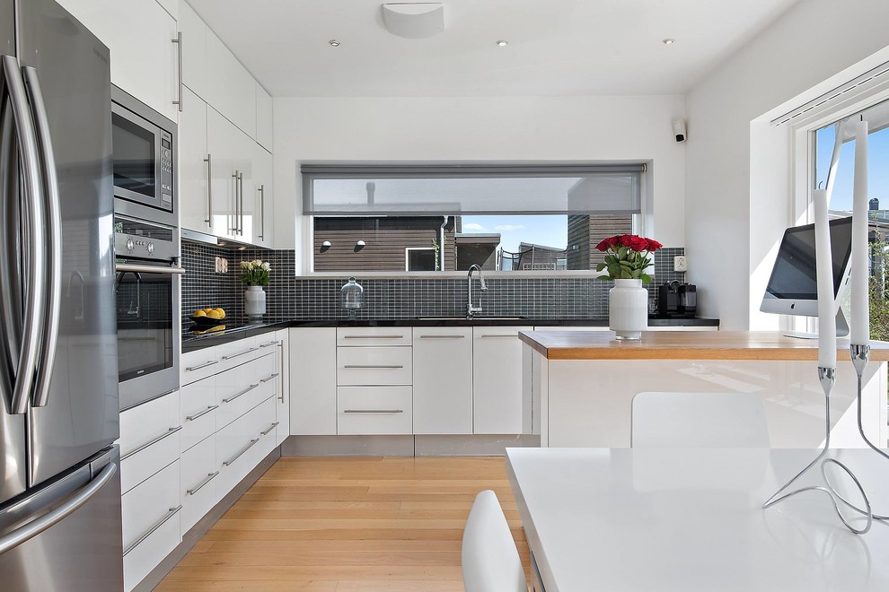 Inspiration for a medium sized classic l-shaped enclosed kitchen in Stockholm with a submerged sink, flat-panel cabinets, white cabinets, black splashback, stainless steel appliances, light hardwood flooring and a breakfast bar.