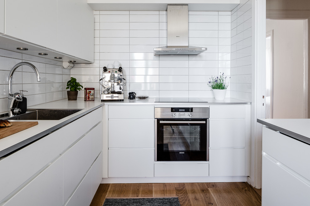 Exempel på ett mellanstort skandinaviskt l-kök, med en undermonterad diskho, släta luckor, vita skåp, vitt stänkskydd, rostfria vitvaror, mörkt trägolv och en köksö