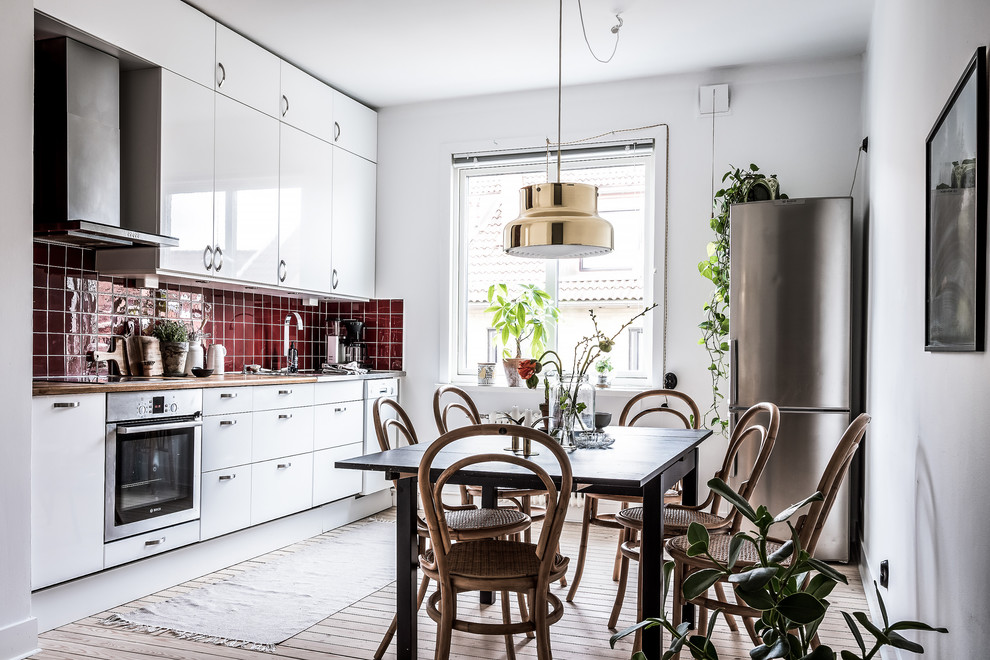 Photo of a scandinavian single-wall kitchen/diner in Gothenburg with flat-panel cabinets, white cabinets, wood worktops, red splashback, light hardwood flooring, no island, beige floors, a built-in sink, ceramic splashback and stainless steel appliances.