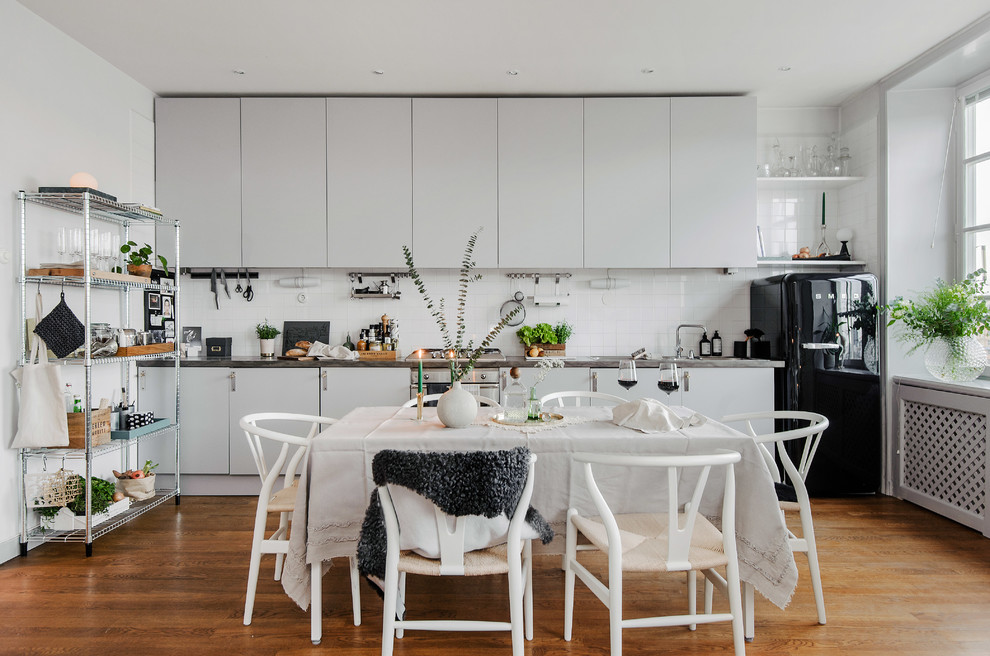 Skandinavisk inredning av ett mellanstort linjärt kök och matrum, med släta luckor, grå skåp, bänkskiva i rostfritt stål, vitt stänkskydd, svarta vitvaror och mellanmörkt trägolv