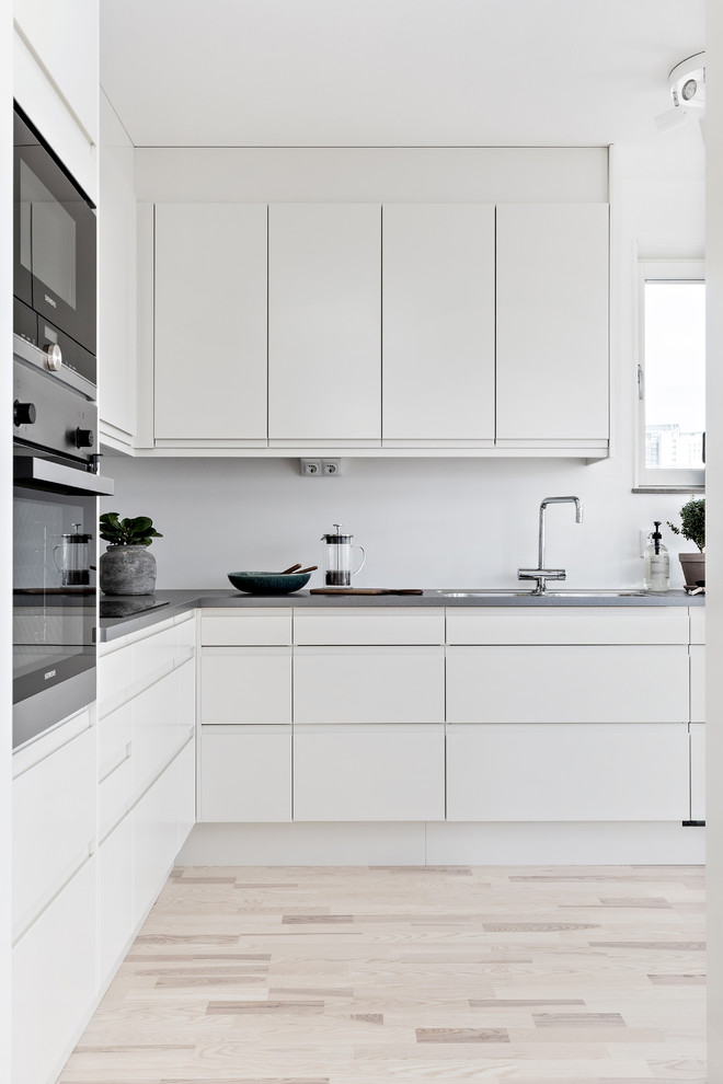 Ejemplo de cocinas en L escandinava con armarios con paneles lisos, puertas de armario blancas, electrodomésticos de acero inoxidable, suelo de madera clara, suelo beige y encimeras grises