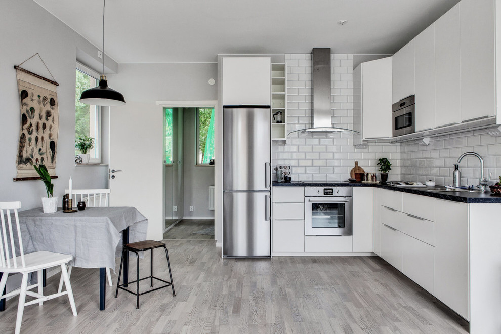 Scandinavian l-shaped kitchen/diner in Stockholm with flat-panel cabinets, white cabinets, white splashback, no island, grey floors, black worktops, a submerged sink, metro tiled splashback, stainless steel appliances and light hardwood flooring.