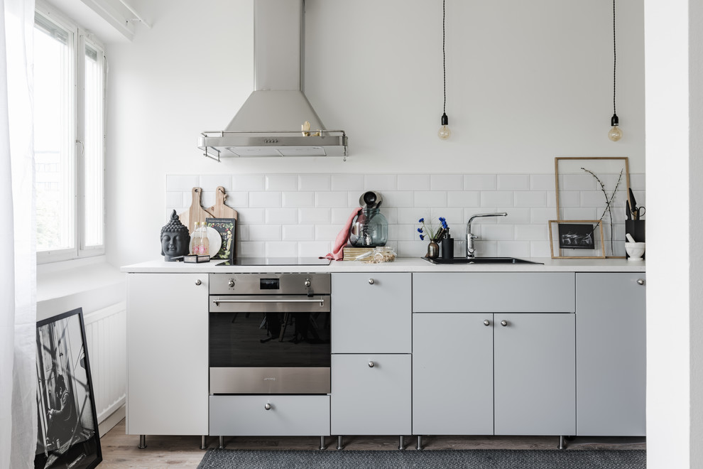 Example of a mid-sized danish single-wall medium tone wood floor enclosed kitchen design in Stockholm with flat-panel cabinets, gray cabinets, no island, a single-bowl sink, white backsplash, ceramic backsplash, stainless steel appliances and white countertops