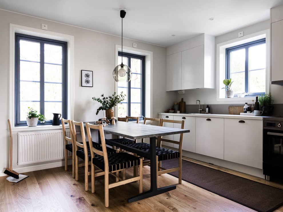 На фото: прямая кухня в скандинавском стиле с обеденным столом, плоскими фасадами, белыми фасадами, черной техникой, паркетным полом среднего тона, коричневым полом и окном без острова с