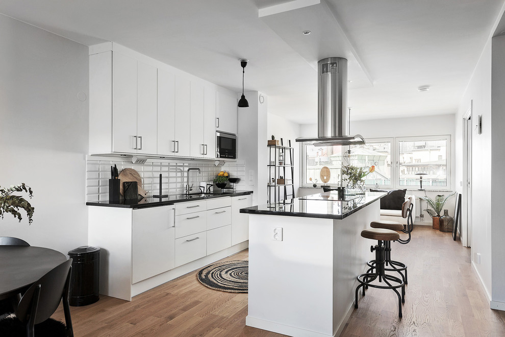 Photo of a medium sized scandinavian galley open plan kitchen in Stockholm with a submerged sink, flat-panel cabinets, white cabinets, white splashback, metro tiled splashback, stainless steel appliances, light hardwood flooring and an island.