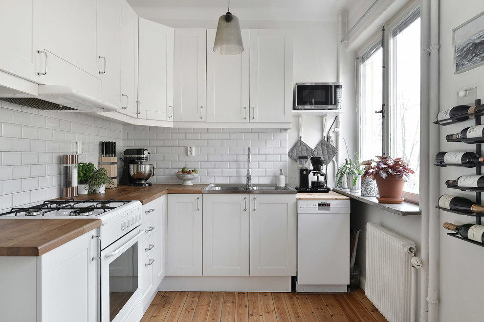 Inspiration for a scandi l-shaped kitchen in Stockholm with a double-bowl sink, shaker cabinets, white cabinets, wood worktops, white splashback, metro tiled splashback, white appliances and light hardwood flooring.