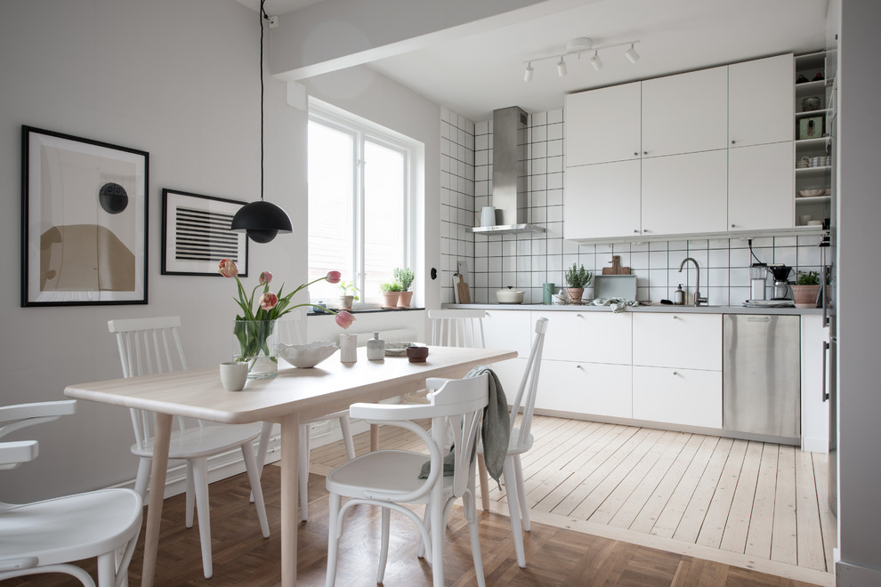 Small scandi single-wall kitchen/diner in Gothenburg with flat-panel cabinets, white cabinets, stainless steel worktops, white splashback, stainless steel appliances, light hardwood flooring, no island and beige floors.