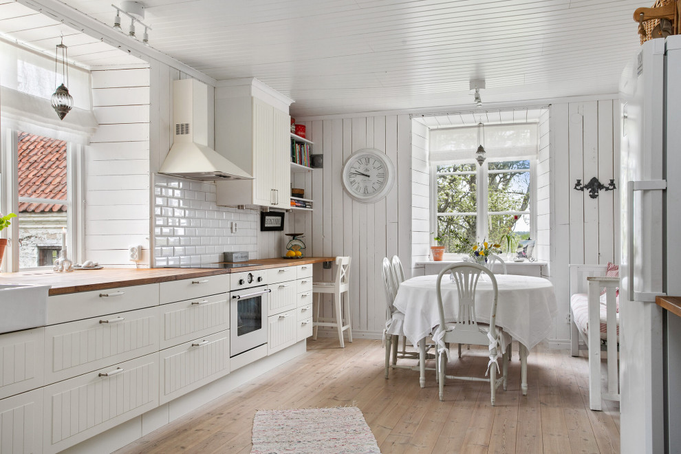 Eat-in kitchen - mid-sized cottage single-wall light wood floor and beige floor eat-in kitchen idea in Other with a farmhouse sink, white cabinets, wood countertops, white backsplash, subway tile backsplash, white appliances, no island, beige countertops and flat-panel cabinets