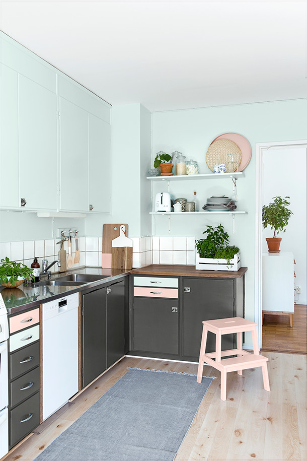 Nordisk inredning av ett litet l-kök, med en dubbel diskho, grå skåp, vitt stänkskydd, vita vitvaror och ljust trägolv