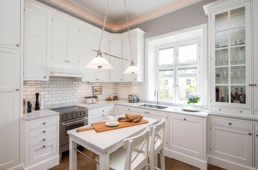 This is an example of a medium sized victorian l-shaped enclosed kitchen in Stockholm with a submerged sink, shaker cabinets, white cabinets, white splashback, metro tiled splashback, an island, marble worktops, stainless steel appliances and medium hardwood flooring.