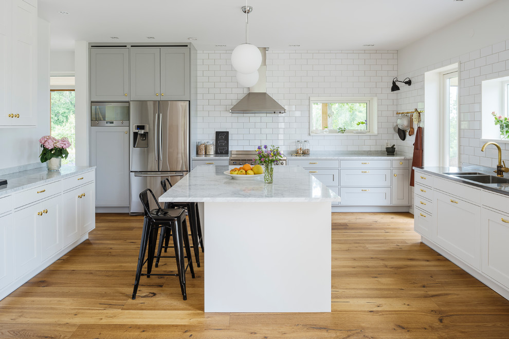 Large traditional kitchen/diner in Gothenburg with a double-bowl sink, shaker cabinets, marble worktops, stainless steel appliances, an island, white cabinets, white splashback, metro tiled splashback and light hardwood flooring.