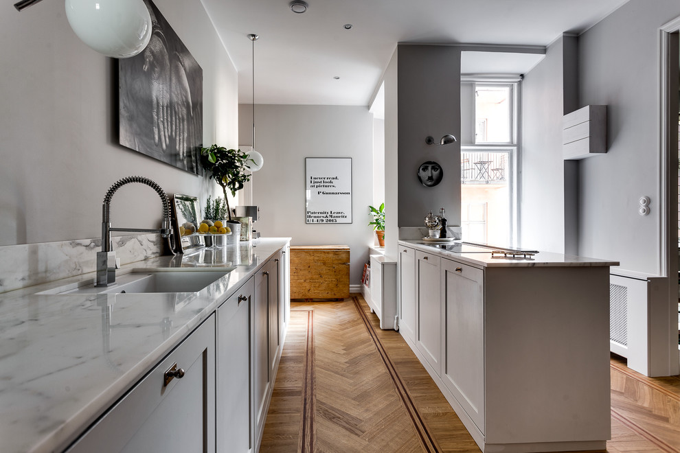 Medium sized scandi galley kitchen in Stockholm with a submerged sink, shaker cabinets, white cabinets, marble worktops, stainless steel appliances, light hardwood flooring and a breakfast bar.