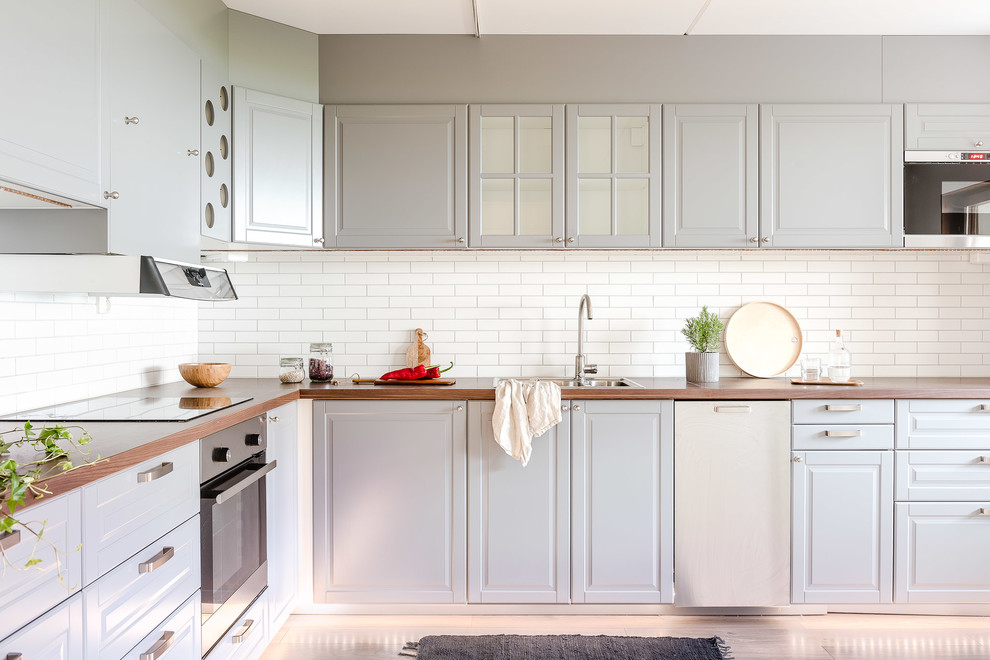 Exempel på ett avskilt, mellanstort skandinaviskt l-kök, med en nedsänkt diskho, vitt stänkskydd, stänkskydd i tunnelbanekakel, rostfria vitvaror, luckor med upphöjd panel, vita skåp och träbänkskiva