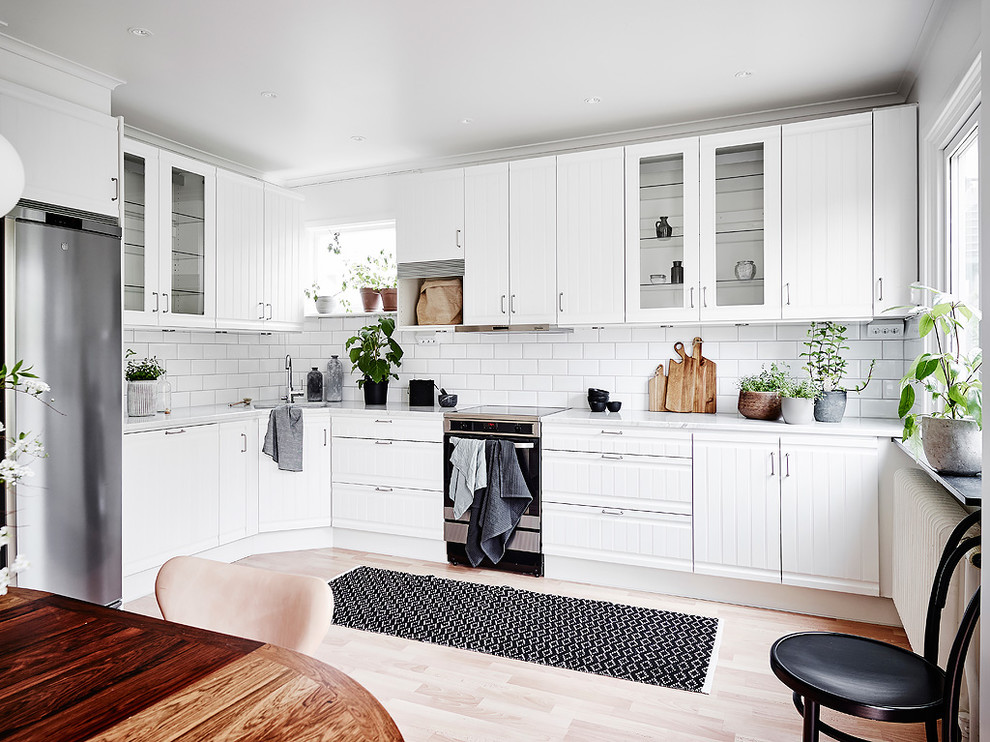Nordische Wohnküche ohne Insel in L-Form mit weißen Schränken, Küchenrückwand in Weiß, Rückwand aus Metrofliesen, Küchengeräten aus Edelstahl, hellem Holzboden und beigem Boden in Göteborg