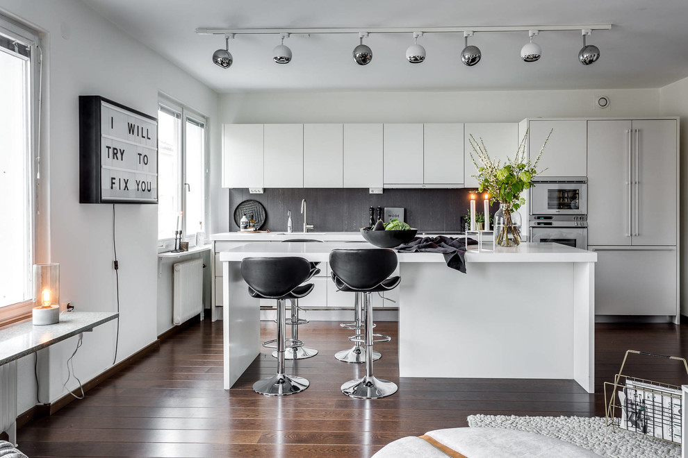 Bild på ett funkis kök med öppen planlösning, med en undermonterad diskho, släta luckor, vita skåp, grått stänkskydd, vita vitvaror, mörkt trägolv och en köksö