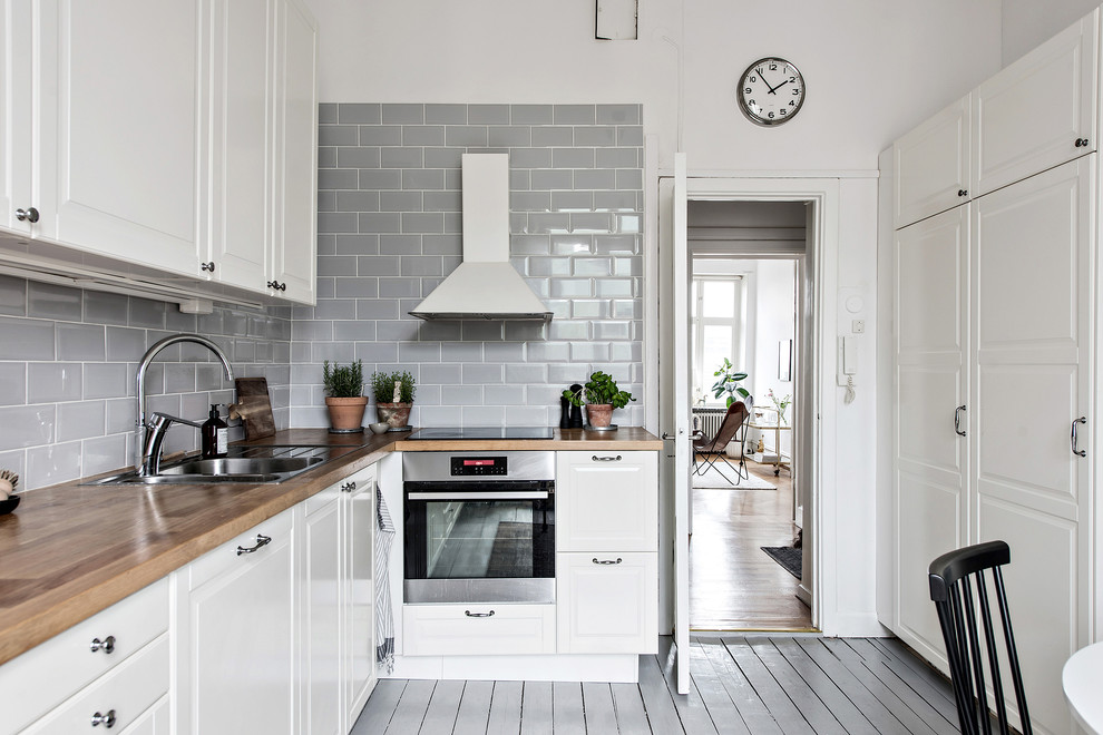Bild på ett litet nordiskt kök, med vita skåp, en dubbel diskho, luckor med upphöjd panel, träbänkskiva, grått stänkskydd, stänkskydd i tunnelbanekakel, målat trägolv och grått golv