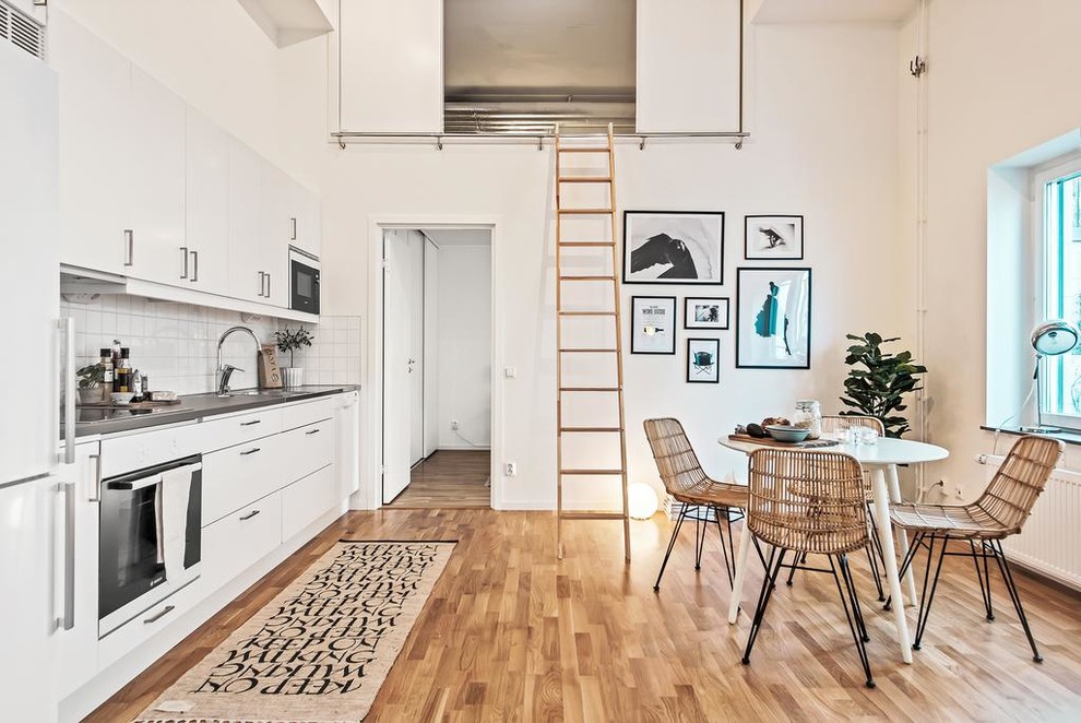 This is an example of a scandi single-wall kitchen/diner in Stockholm with flat-panel cabinets, white cabinets, white splashback, white appliances, light hardwood flooring, no island, beige floors and grey worktops.