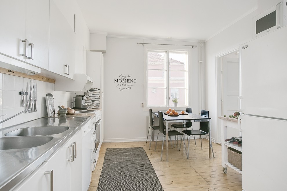 Bild på ett mellanstort nordiskt linjärt kök och matrum, med en integrerad diskho, släta luckor, vita skåp, bänkskiva i rostfritt stål, vitt stänkskydd, vita vitvaror, ljust trägolv och en köksö