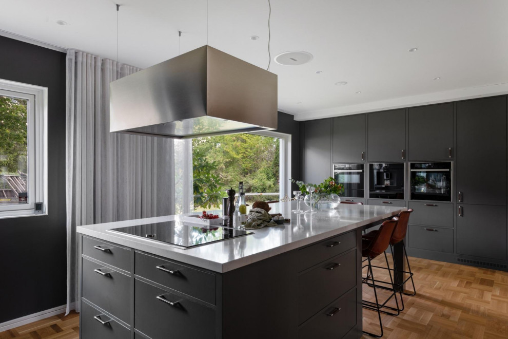 Klassisk inredning av ett grå grått kök, med släta luckor, svarta skåp, svarta vitvaror, mellanmörkt trägolv, en köksö och brunt golv