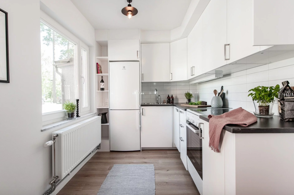Inspiration for a small scandinavian l-shaped open plan kitchen in Stockholm with a double-bowl sink, flat-panel cabinets, white cabinets, laminate countertops, white splashback, porcelain splashback, white appliances, light hardwood flooring, beige floors and grey worktops.