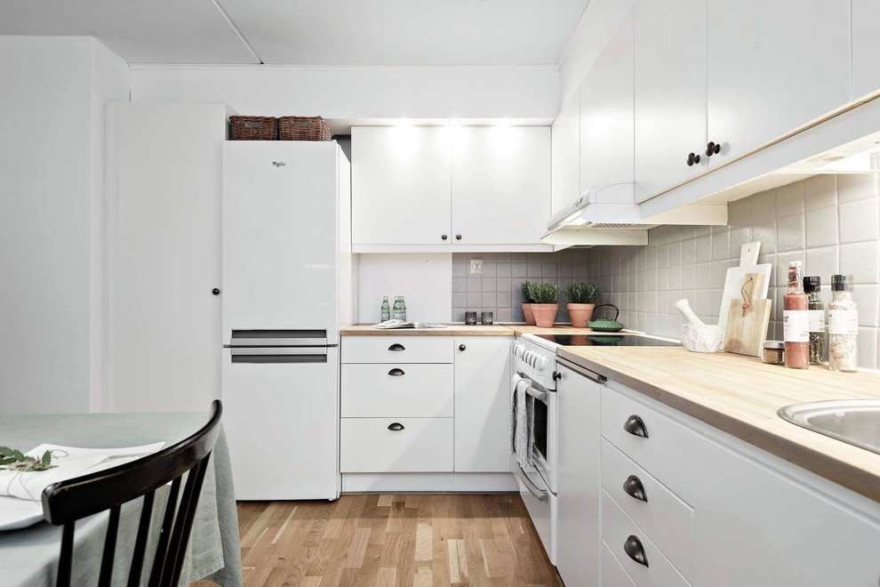 This is an example of a scandinavian l-shaped kitchen/diner in Stockholm with a built-in sink, flat-panel cabinets, white cabinets, wood worktops, grey splashback, white appliances, medium hardwood flooring, brown floors and beige worktops.