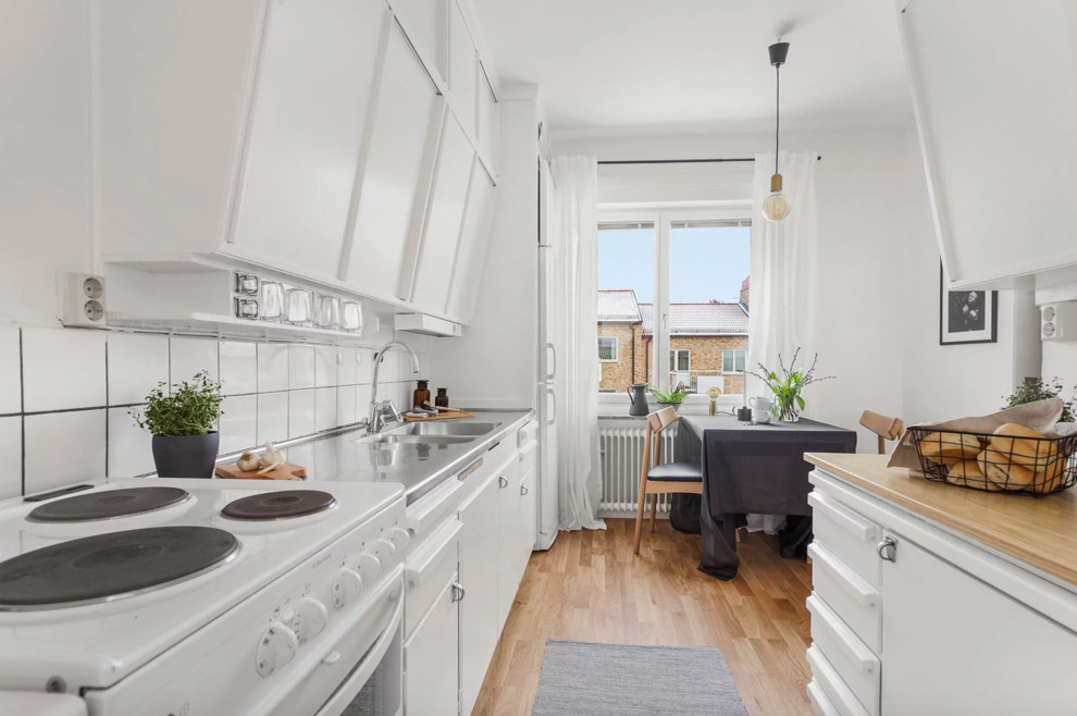 Bild på ett mellanstort nordiskt kök, med en dubbel diskho, släta luckor, vita skåp, bänkskiva i rostfritt stål, vitt stänkskydd, stänkskydd i porslinskakel, vita vitvaror, ljust trägolv och beiget golv