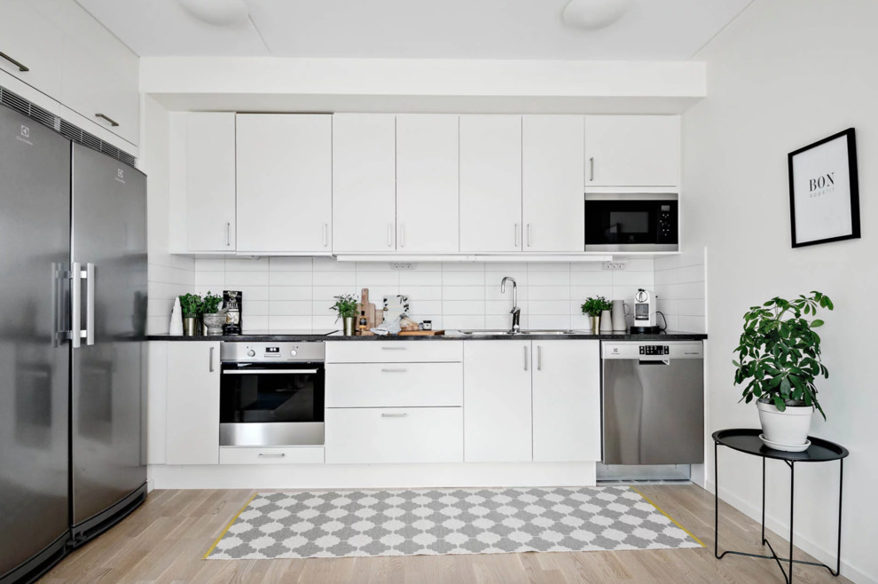 Foto på ett litet minimalistiskt linjärt kök med öppen planlösning, med en dubbel diskho, släta luckor, vita skåp, laminatbänkskiva, vitt stänkskydd, stänkskydd i porslinskakel, rostfria vitvaror, ljust trägolv och beiget golv
