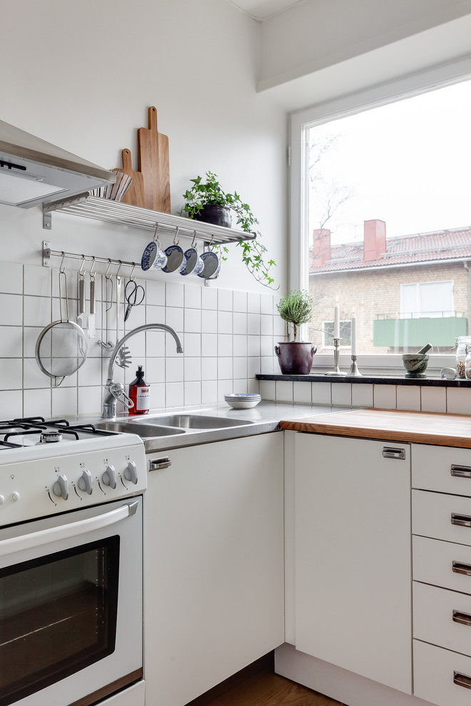 На фото: отдельная, угловая кухня среднего размера в скандинавском стиле с двойной мойкой, плоскими фасадами, белыми фасадами, деревянной столешницей, белым фартуком и белой техникой без острова