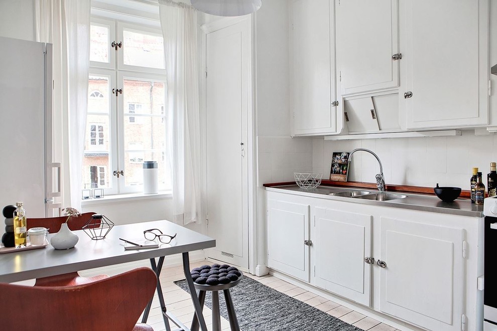 Exempel på ett minimalistiskt linjärt kök och matrum, med en dubbel diskho, luckor med upphöjd panel, vita skåp, bänkskiva i rostfritt stål, vitt stänkskydd, vita vitvaror, ljust trägolv och beiget golv