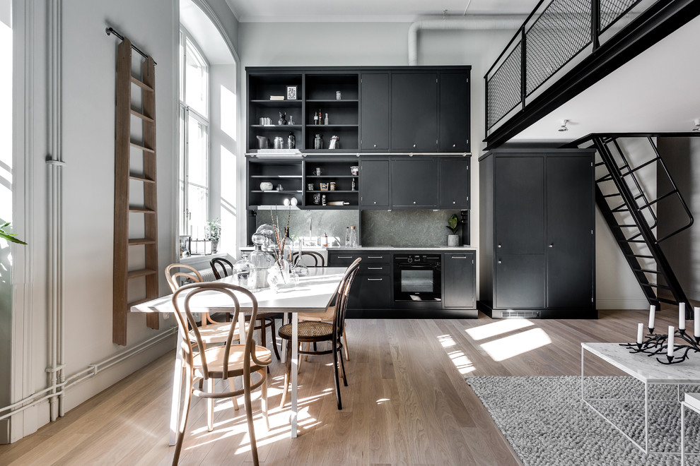 Example of a mid-sized danish single-wall light wood floor open concept kitchen design in Stockholm with limestone countertops, a farmhouse sink, flat-panel cabinets, black cabinets, gray backsplash, stone slab backsplash, black appliances and no island