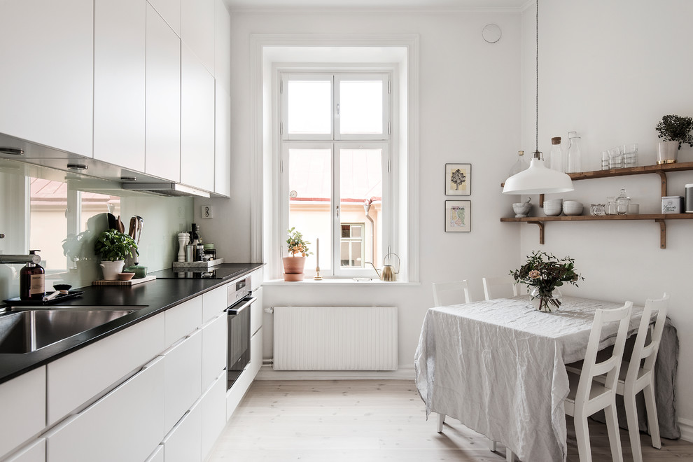 Inredning av ett skandinaviskt linjärt kök, med en undermonterad diskho, släta luckor, vita skåp, grönt stänkskydd, rostfria vitvaror, ljust trägolv och beiget golv