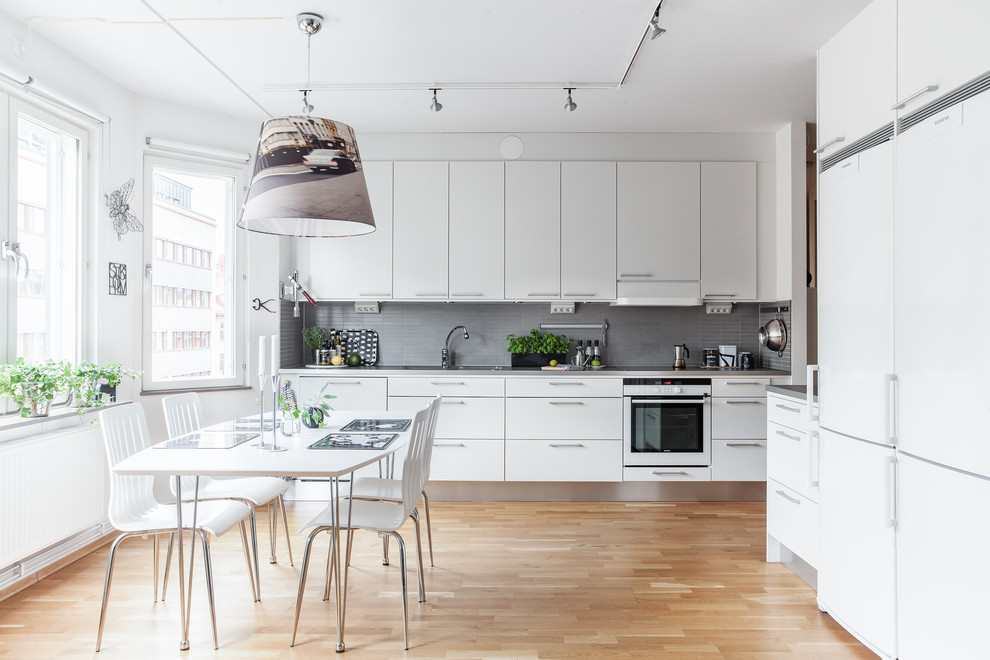 Inspiration for a medium sized scandi l-shaped kitchen/diner in Stockholm with flat-panel cabinets, white cabinets, grey splashback, mosaic tiled splashback, medium hardwood flooring, white appliances and no island.