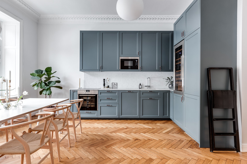Photo of a medium sized scandinavian l-shaped kitchen/diner in Stockholm with blue cabinets, marble splashback, no island, a submerged sink, shaker cabinets, white splashback, stainless steel appliances and light hardwood flooring.