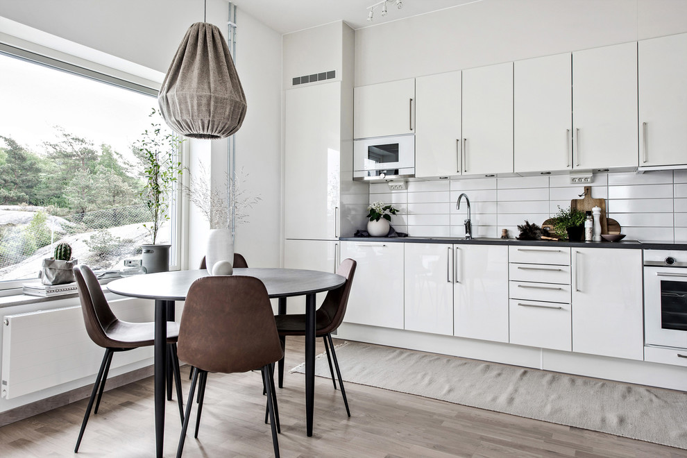 Design ideas for a medium sized scandi single-wall kitchen/diner in Gothenburg with flat-panel cabinets, white cabinets, white splashback, no island, black worktops, light hardwood flooring and beige floors.
