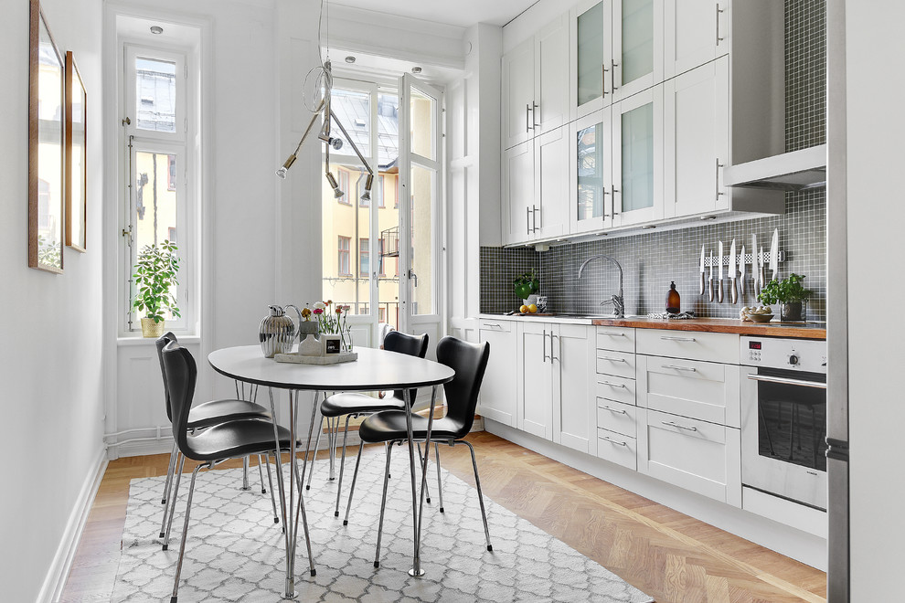 Skandinavisk inredning av ett stort linjärt kök och matrum, med vita skåp, träbänkskiva, stänkskydd i mosaik, vita vitvaror, svart stänkskydd, ljust trägolv och skåp i shakerstil